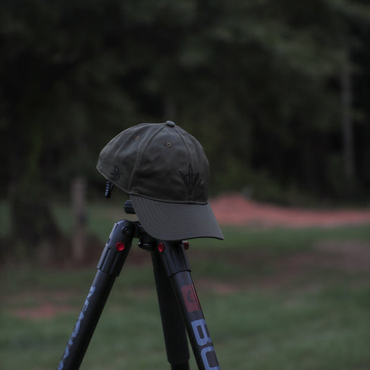 A dark ranger green baseball cap sits on top of a black tripod leg in an outdoor grassy area with a blurred forest background.