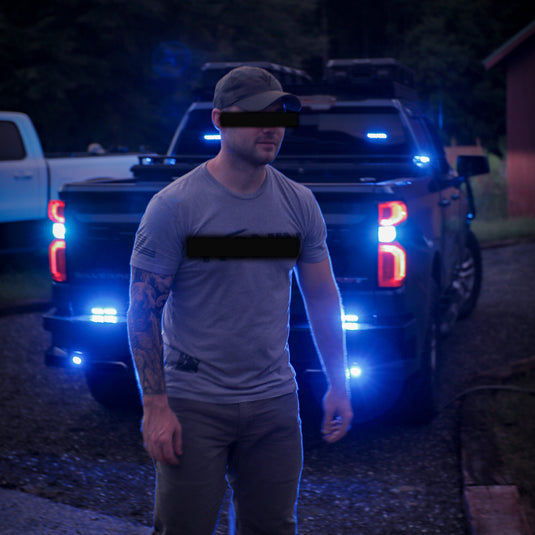 A man wearing a gray t-shirt and dark pants walks in front of a black pickup truck at dusk, with bright blue underglow lights illuminating the scene. The man's face is obscured by a black bar, and there is a white truck partially visible on the left.