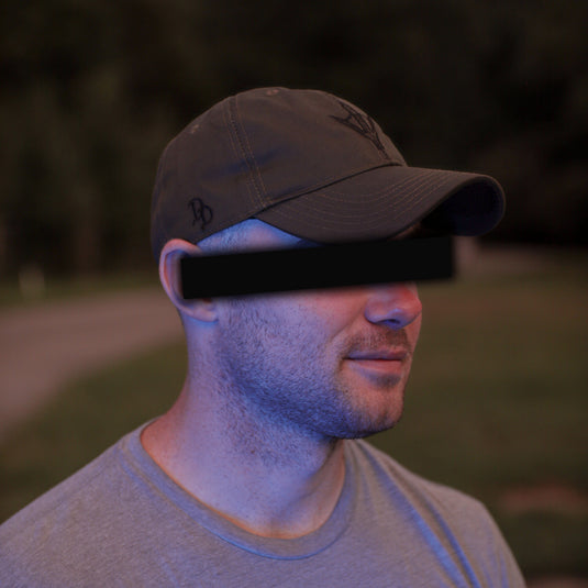 A man wearing a ranger green baseball cap and a gray t-shirt, photographed outdoors at dusk with his eyes obscured by a black bar.