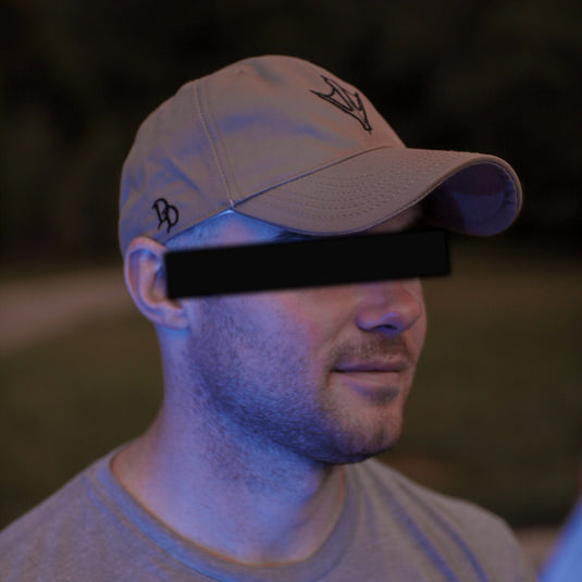 A man wearing a khaki baseball cap with a black embroidered logo and a gray t-shirt, photographed outdoors at dusk with his eyes obscured by a black bar.