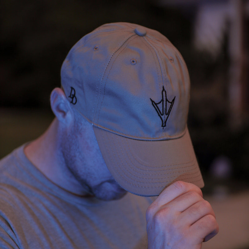 Load image into Gallery viewer, A man wearing a beige baseball cap with a black trident logo and a gray t-shirt, photographed in low light with a strong blue/purple color cast.
