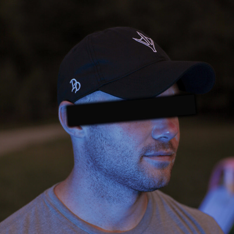 Load image into Gallery viewer, A man wearing a black baseball cap with a white embroidered logo and a gray t-shirt, photographed outdoors at dusk with his eyes obscured by a black bar.
