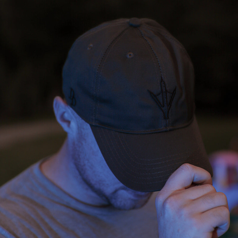 Load image into Gallery viewer, A man wearing a ranger green baseball cap with a black arrowhead logo and a gray t-shirt, photographed in low light with a blue tint against a dark outdoor background.
