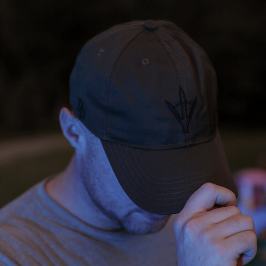 A man wearing a ranger green baseball cap with a black arrowhead logo and a gray t-shirt, photographed in low light with a blue tint against a dark outdoor background.