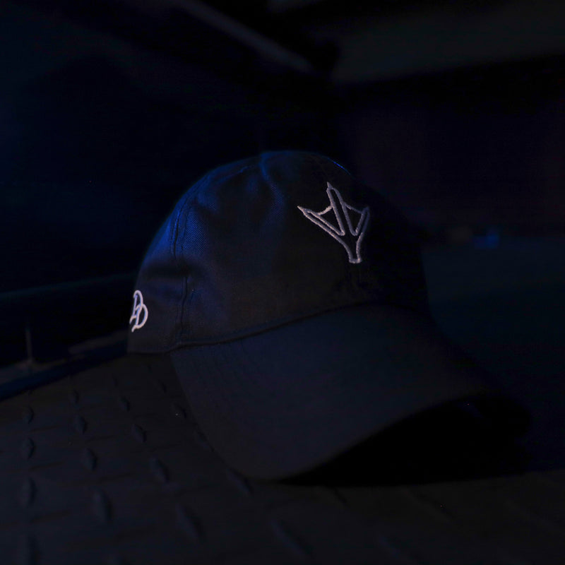 Load image into Gallery viewer, A black baseball cap with white embroidered logos, resting on a textured dark surface in a low-light setting.

